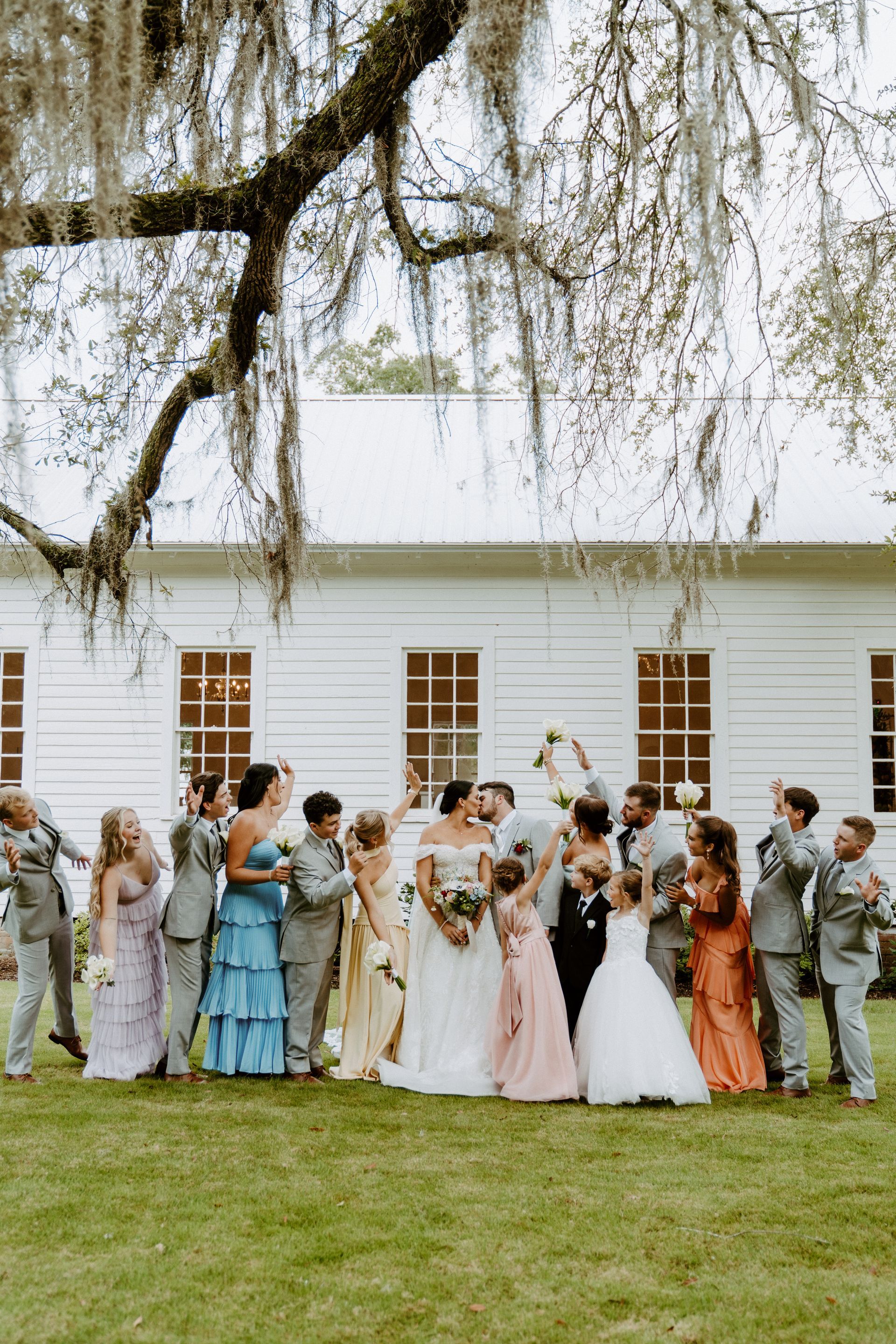 Wedding party posing outside a white building. Bride and groom kiss surrounded by bridesmaids, groomsmen, and children.