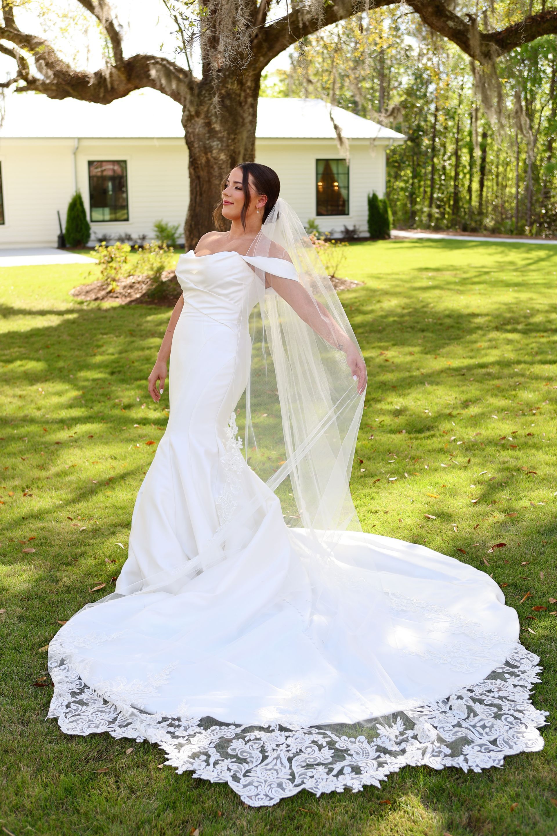 Bride in a white off-the-shoulder gown with a long train poses outdoors in front of a white building.