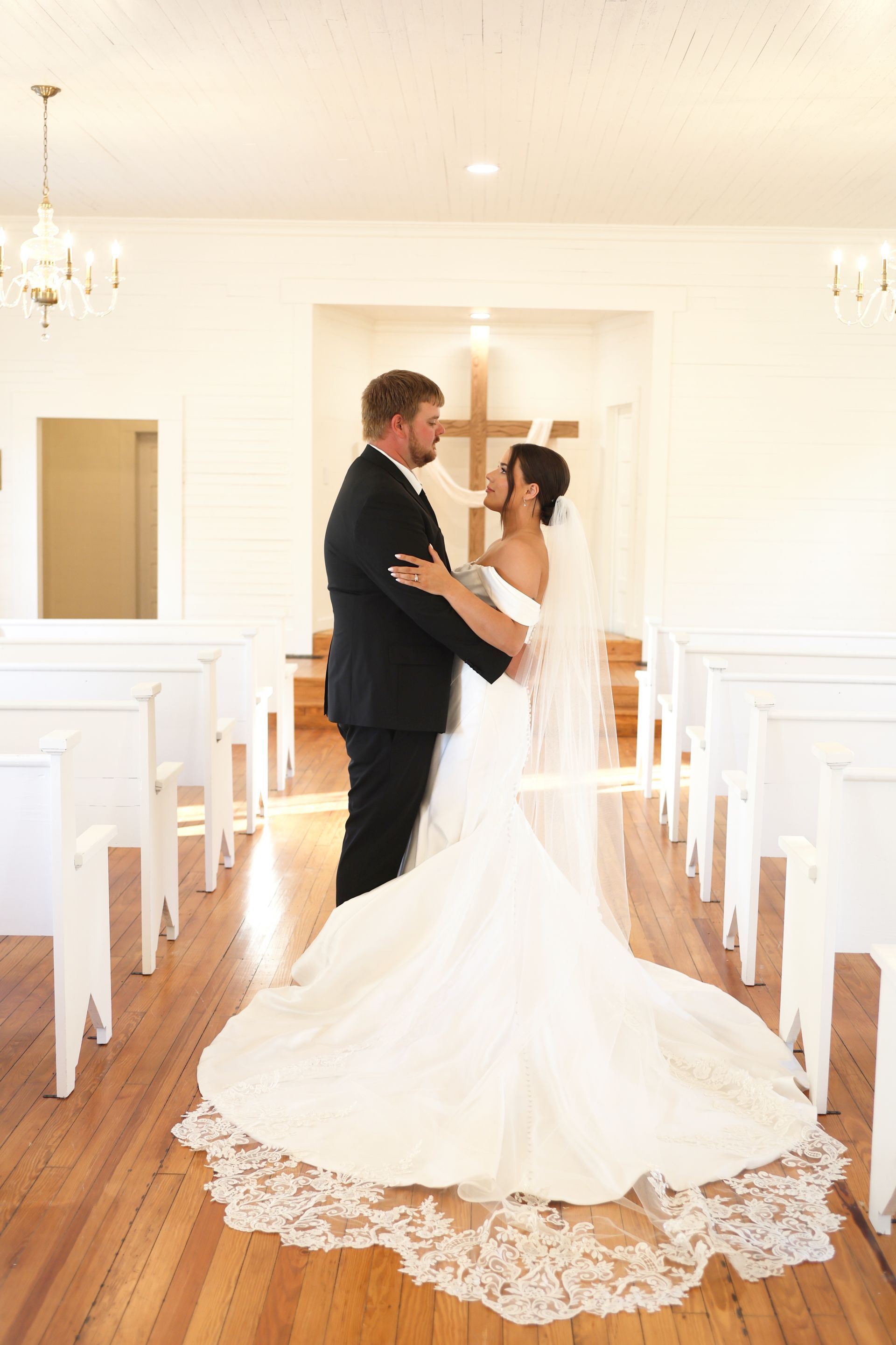 Wedding couple in a white chapel, bride in a gown, groom in a suit, gazing at each other.