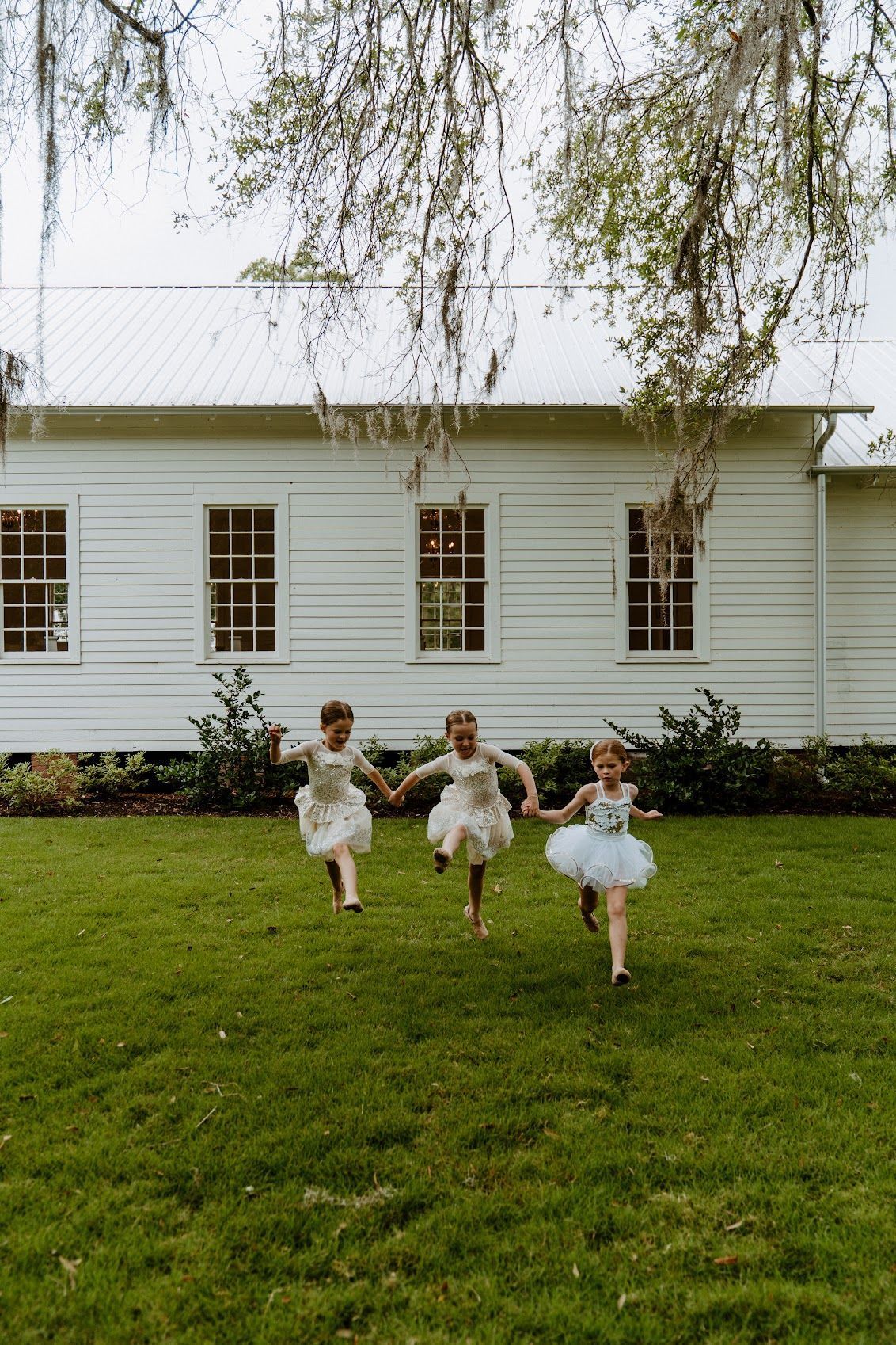 Three young girls in dresses running on a lawn toward a white building.