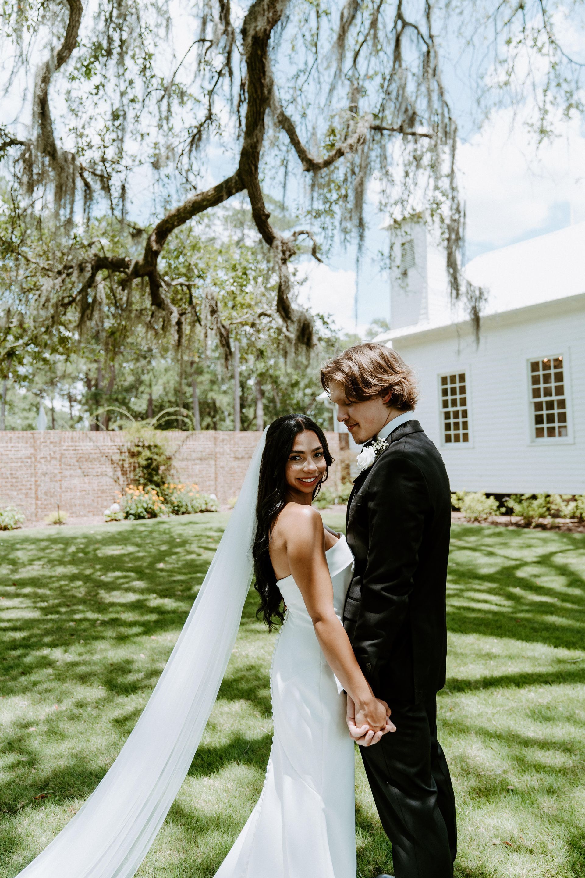 Bride and groom holding hands, smiling at camera, in front of a white building, under tree.