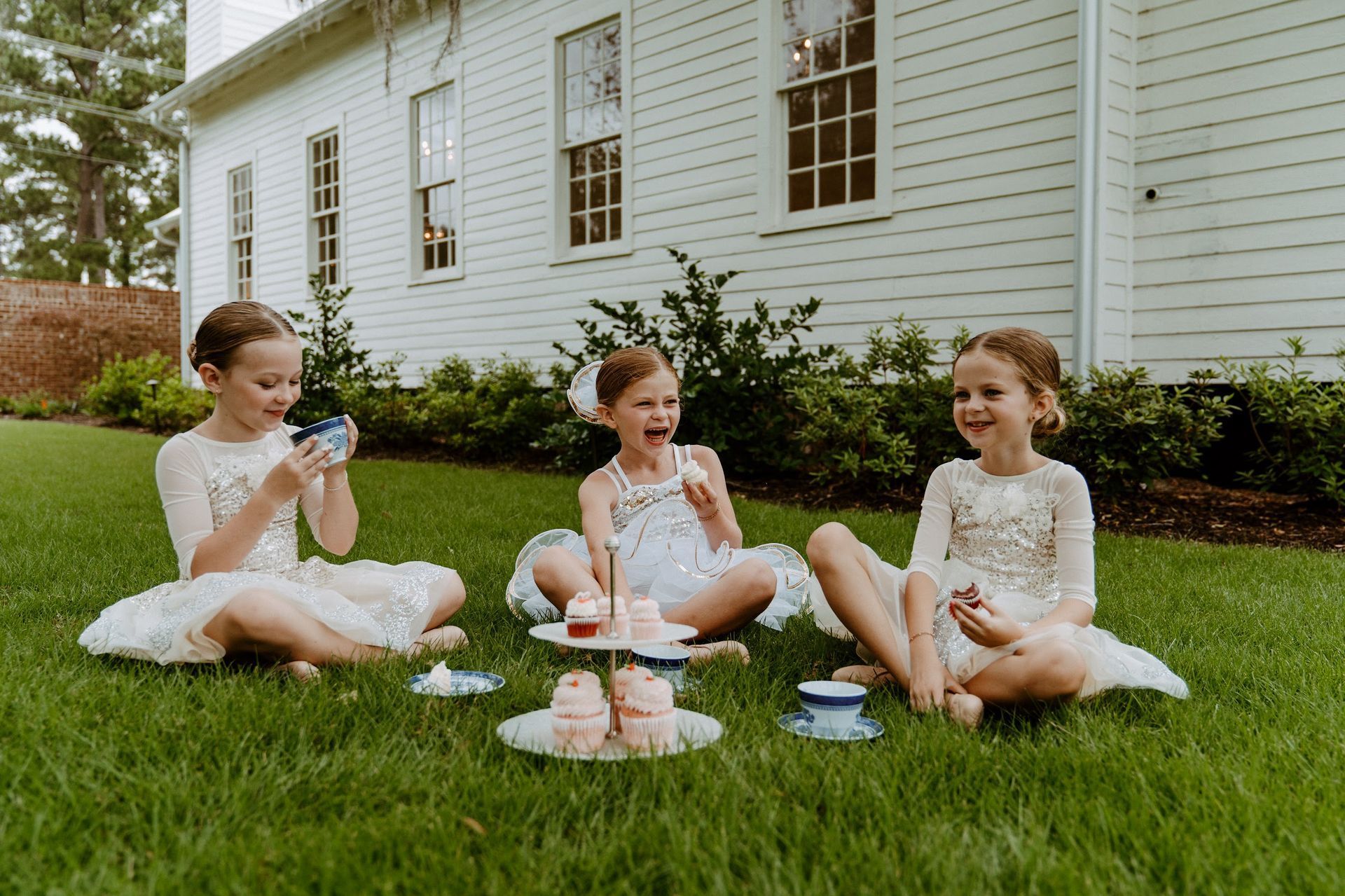 Three girls in white dresses having a tea party on the grass in front of a white house.
