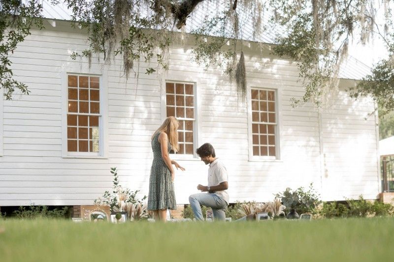 A man is kneeling down to propose to a woman in front of a white house.