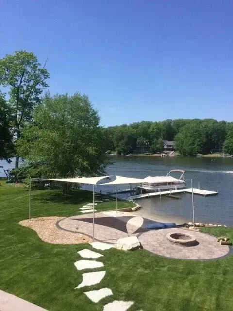 Lakeside scene: boat dock with covered slip, patio area with stone path, green grass, and trees under a clear blue sky.