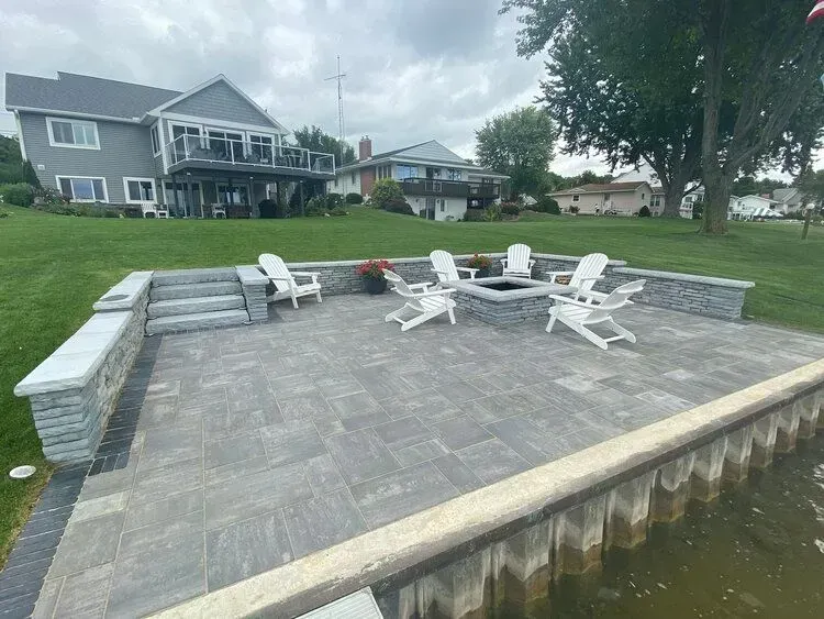 Patio with fire pit and chairs near water, with houses in the background. Gray and white tones.