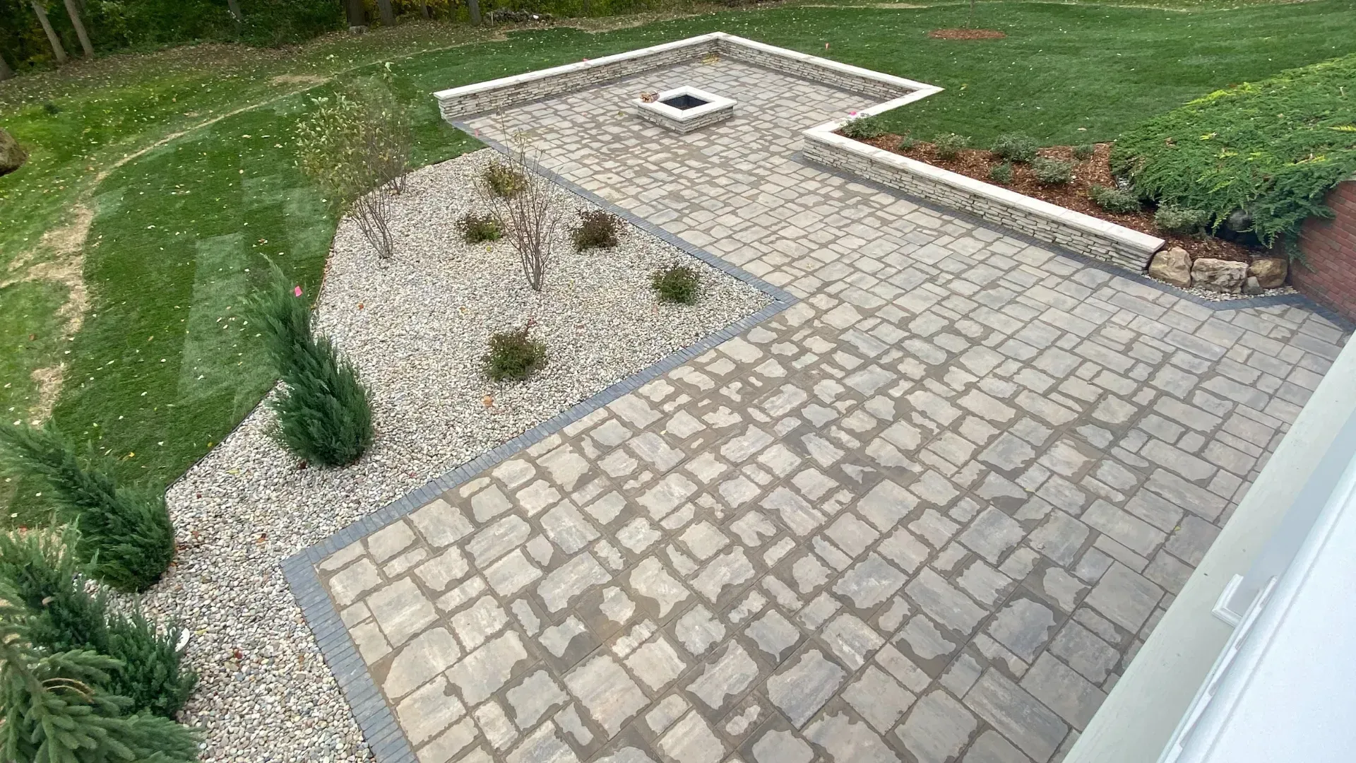Stone patio with fire pit, bordered by gravel and retaining walls, leading to a grassy yard.