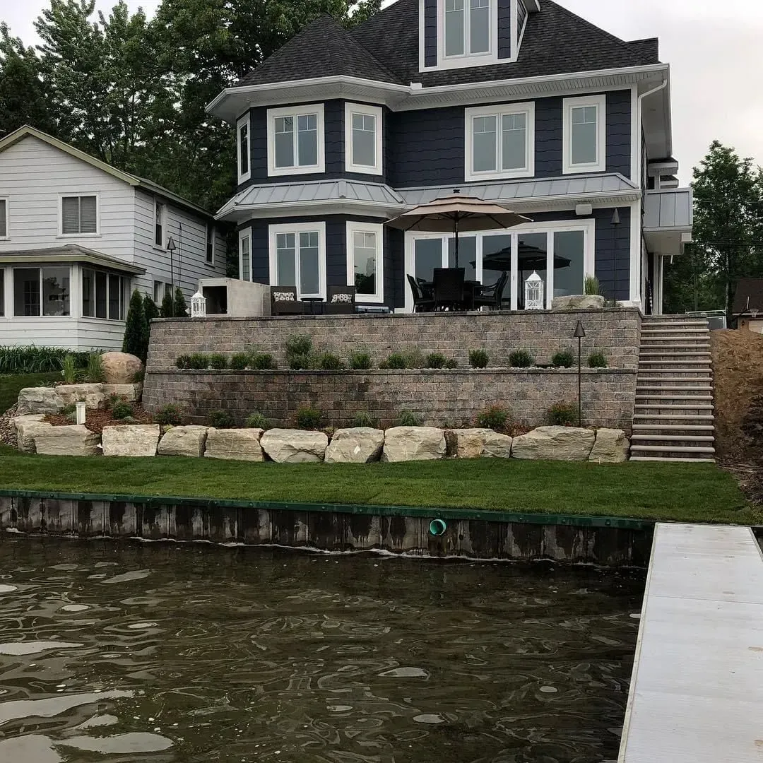 Two-story dark blue house on a lake with stone retaining walls, patio, and stairs.