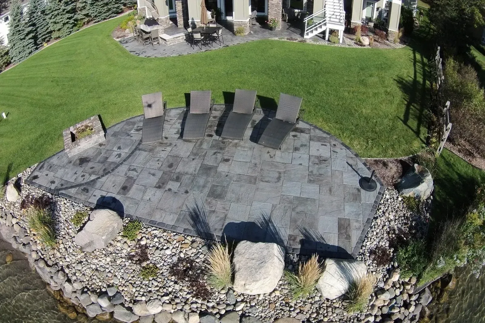 Overhead view of a stone patio with lounge chairs on a grassy lawn near water.