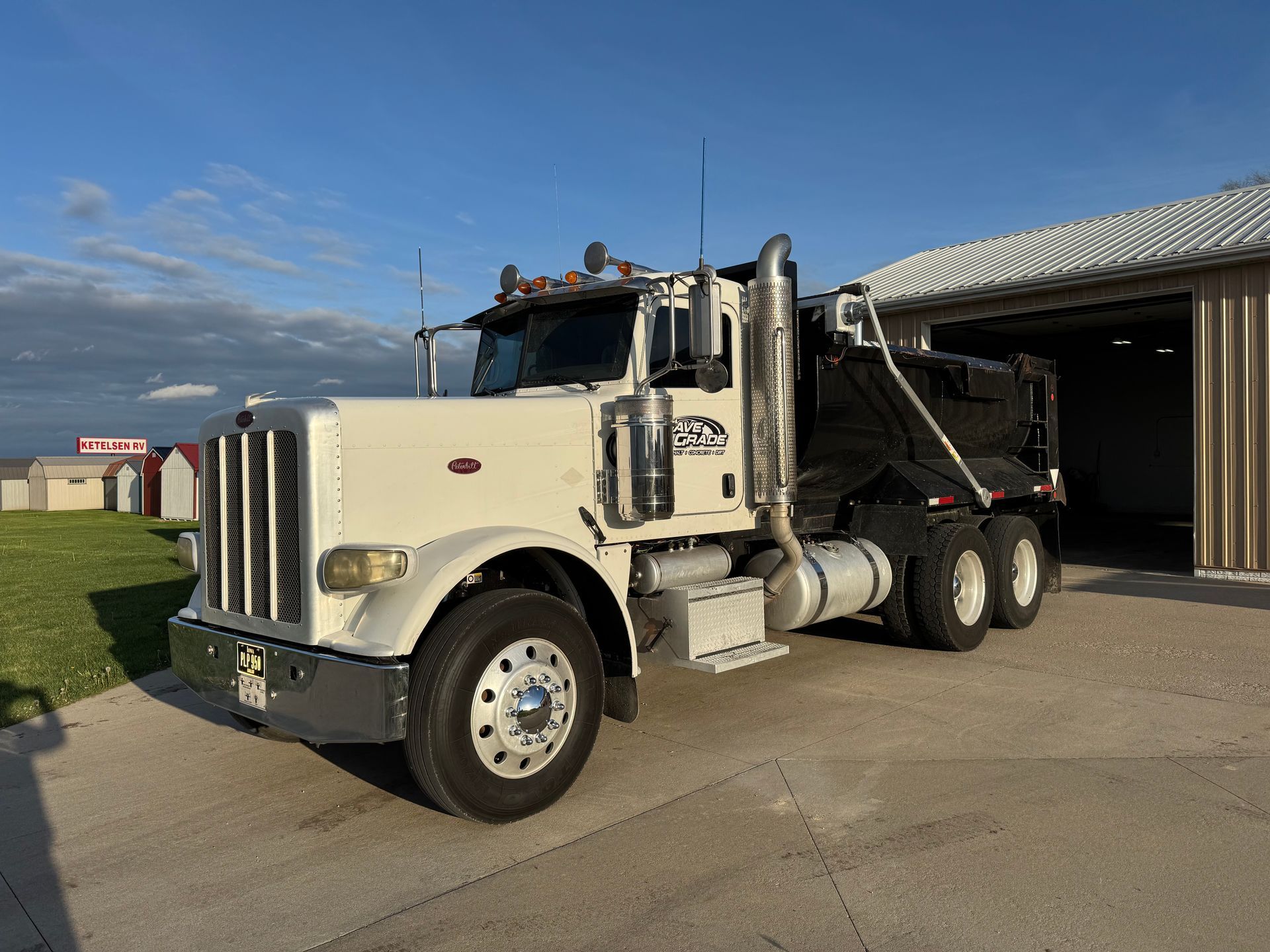 A white Peterbilt dump truck parked on a concrete surface outside a shed under a blue sky.