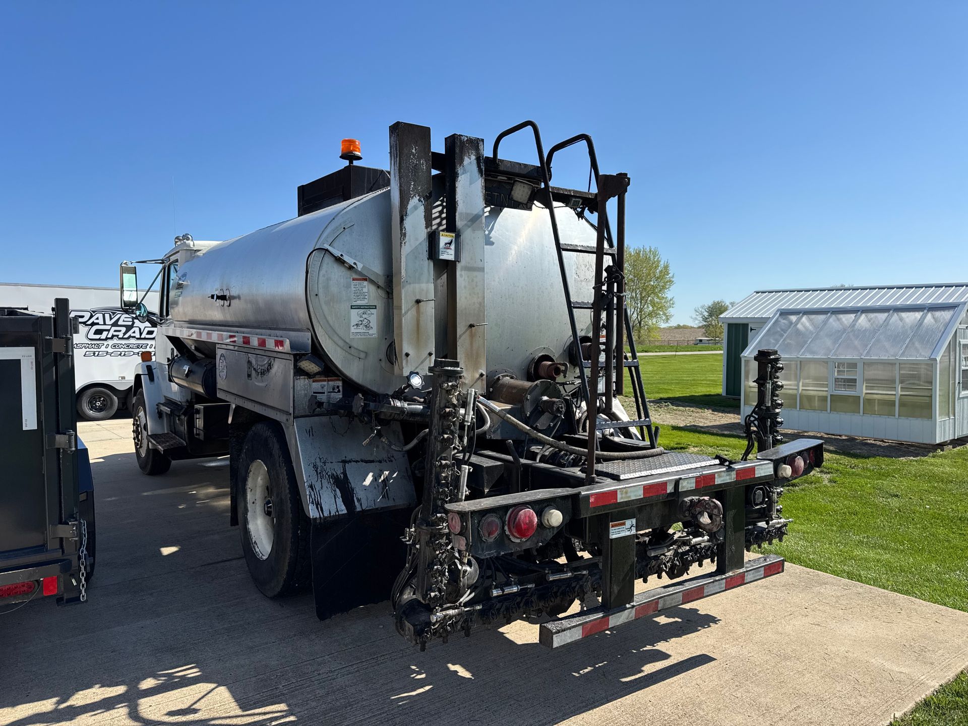 A silver tanker truck for road construction parked on a gravel lot next to a greenhouse.