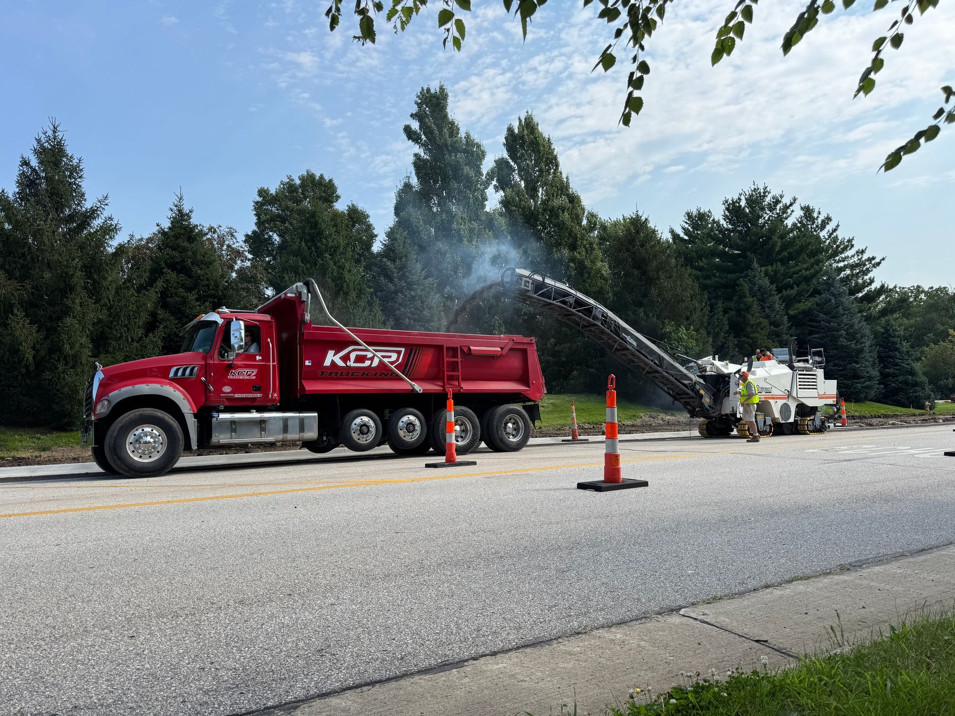A red K-Five dump truck is being loaded with asphalt by a milling machine on a suburban road under a sunny sky.