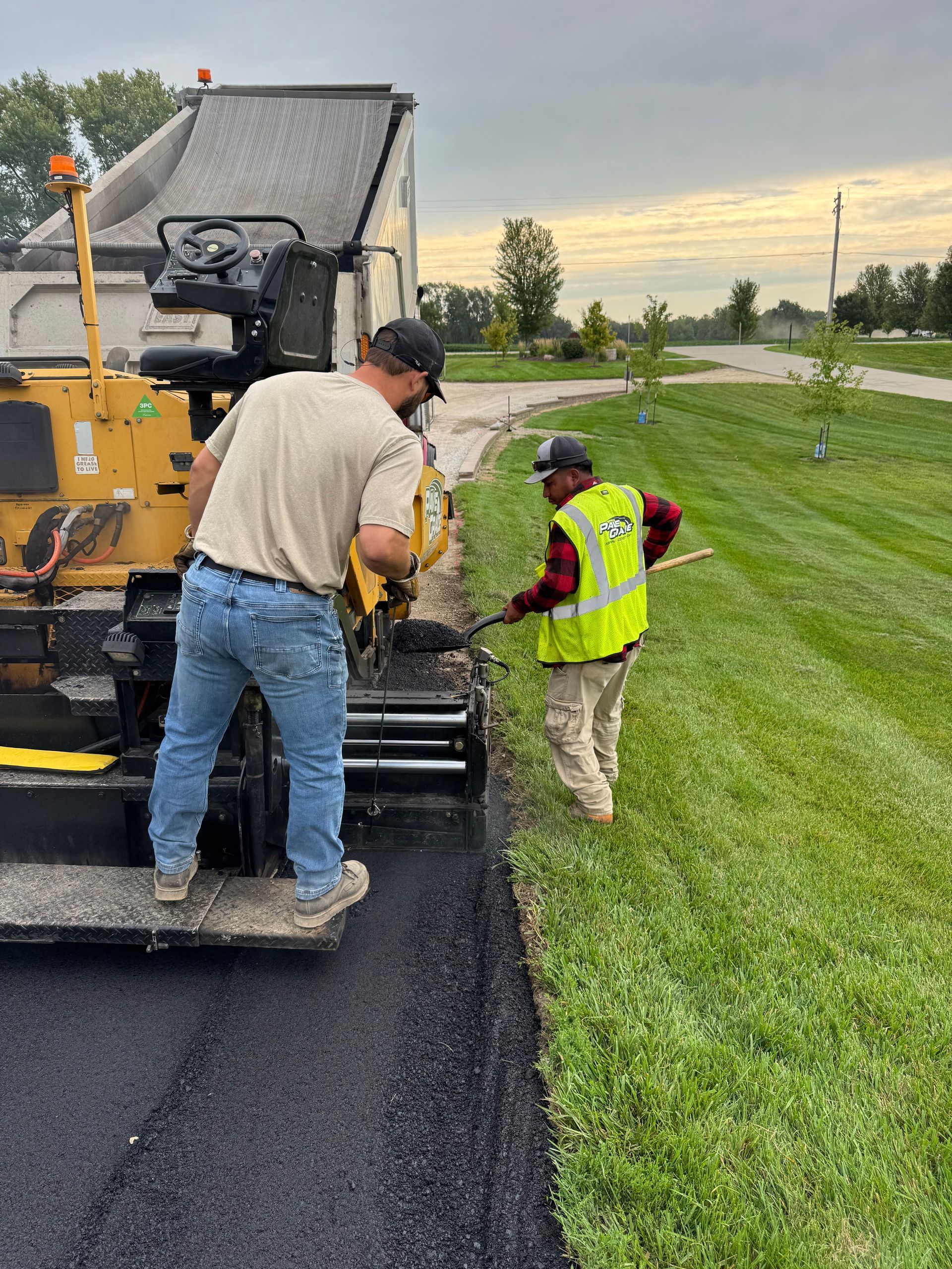 Two construction workers operate an asphalt paving machine to lay a new road surface next to a grassy roadside.