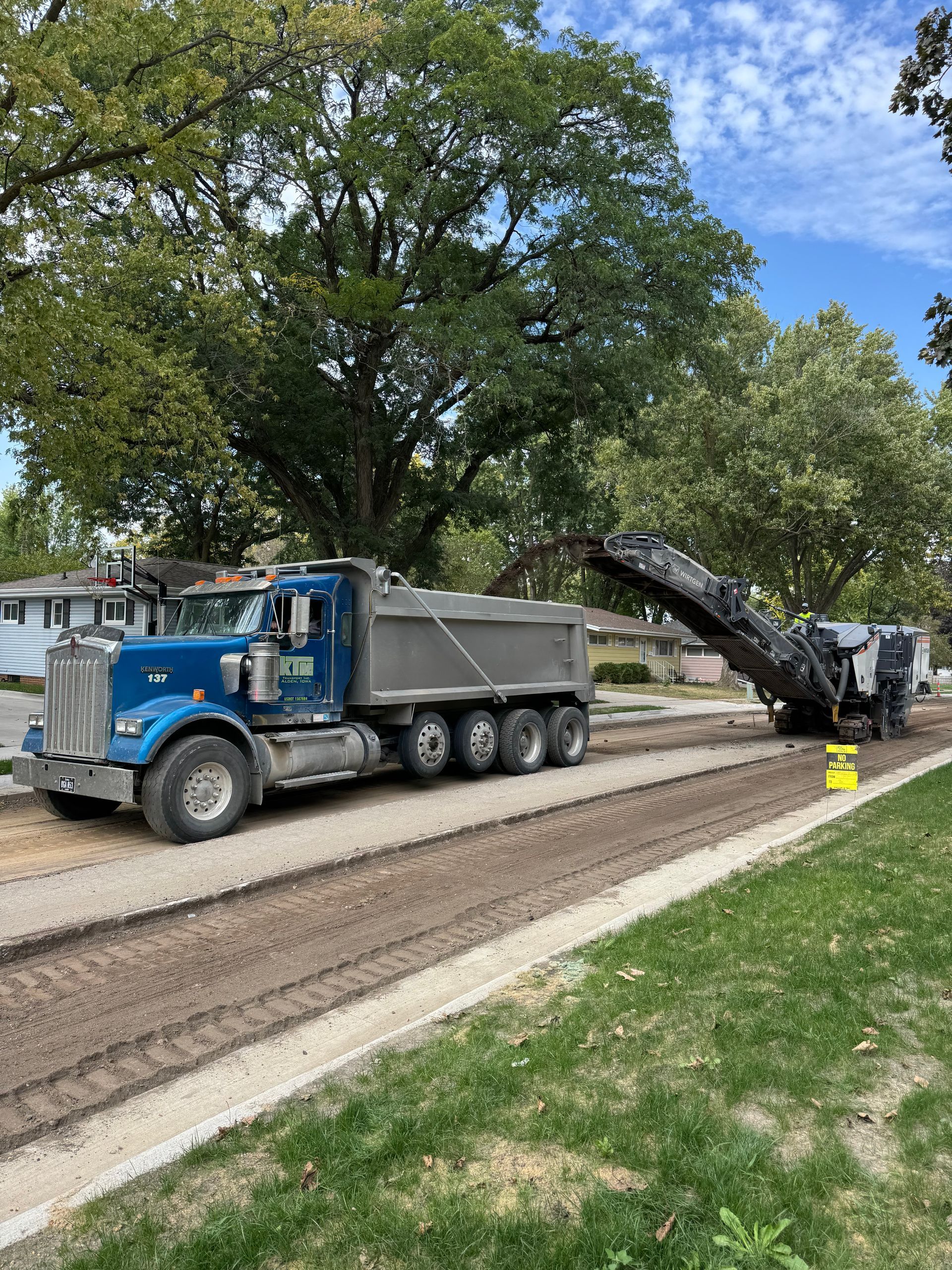 A blue dump truck is parked on a street being resurfaced, loading debris from a milling machine in a suburban setting.
