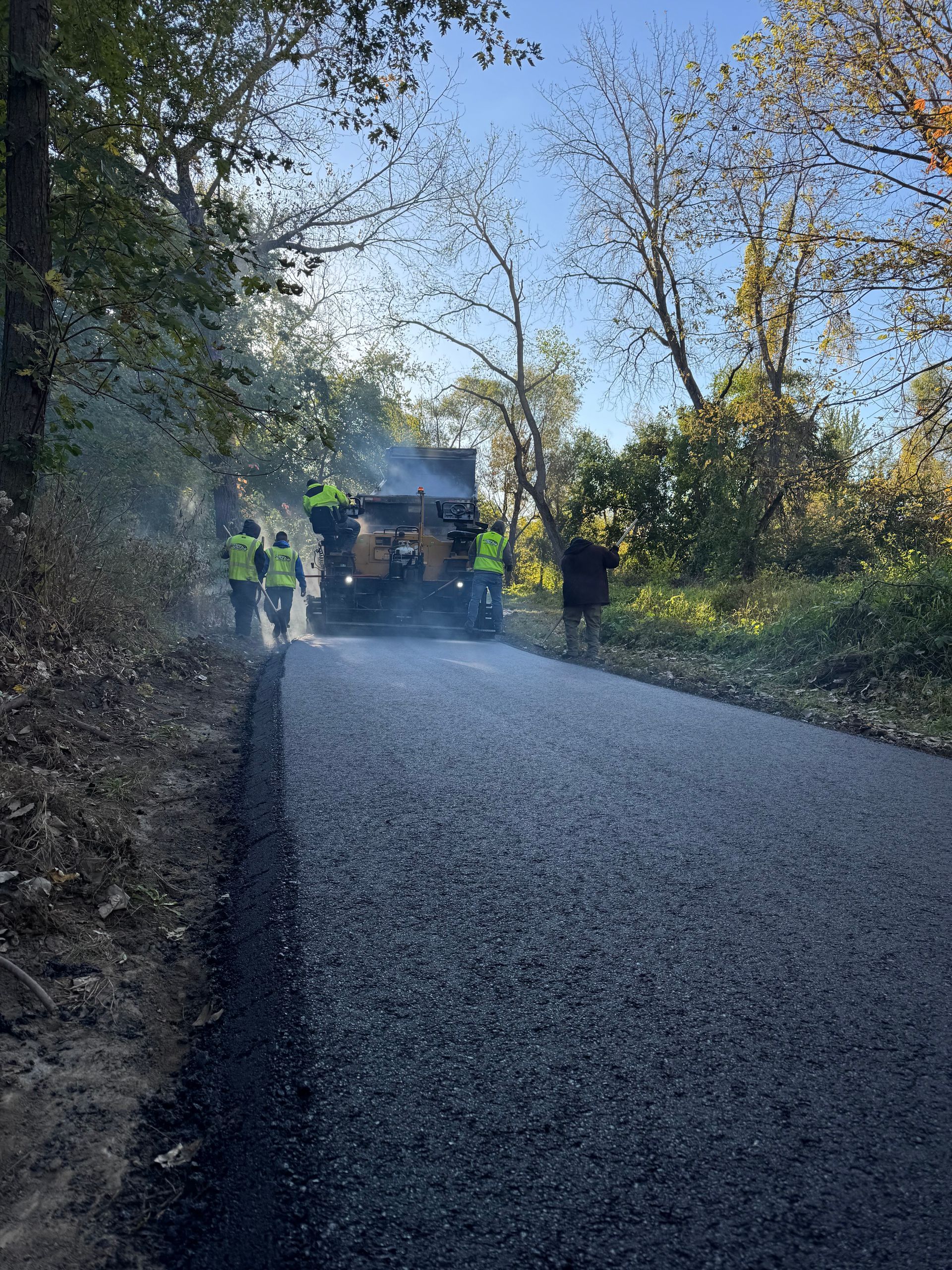 Construction workers in high-visibility vests pave a fresh asphalt road in a wooded area on a sunny day.