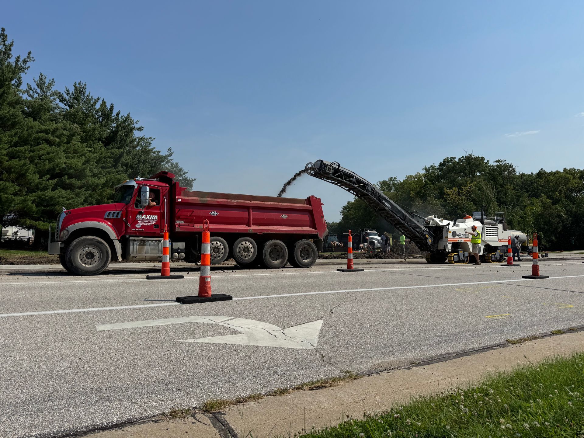 A red dump truck is being loaded with asphalt debris by a road milling machine on a sunny day with orange traffic cones.