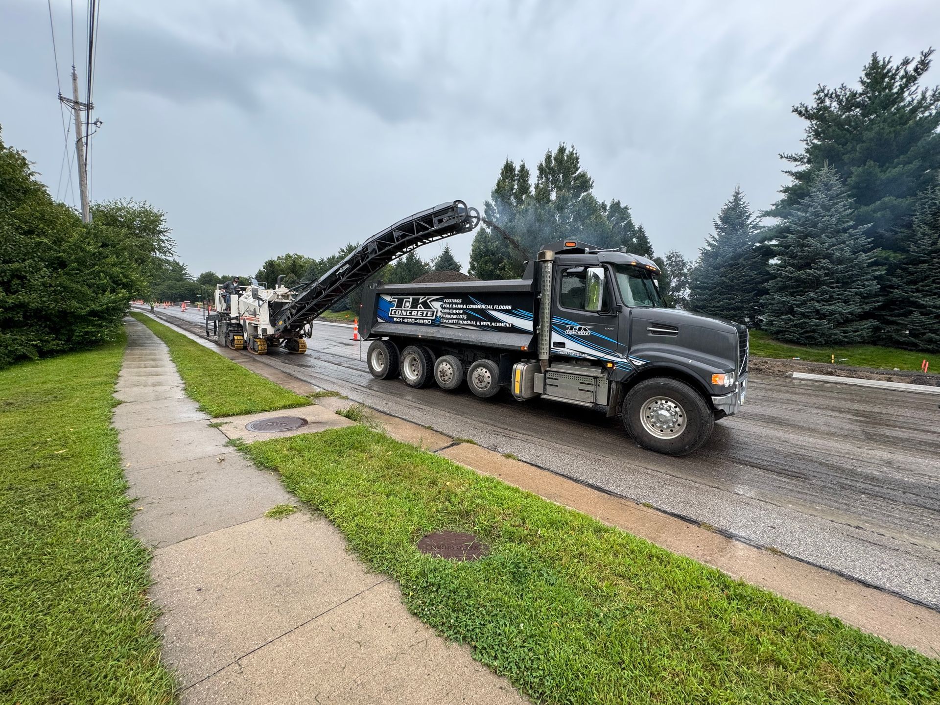 A dark-colored dump truck being loaded by a road milling machine on a suburban street.