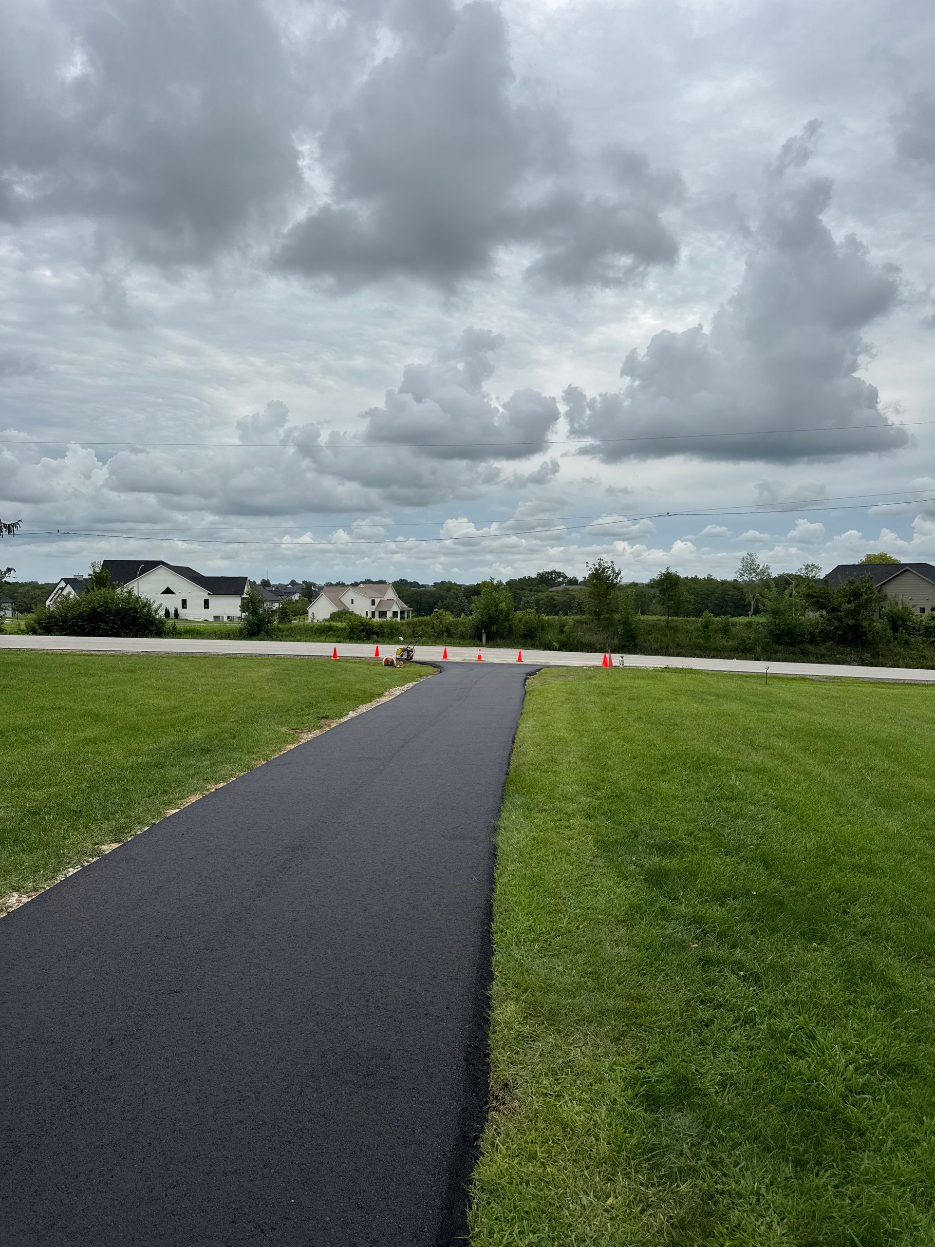 A paved asphalt path leads through a grassy field toward a road under a cloudy sky.