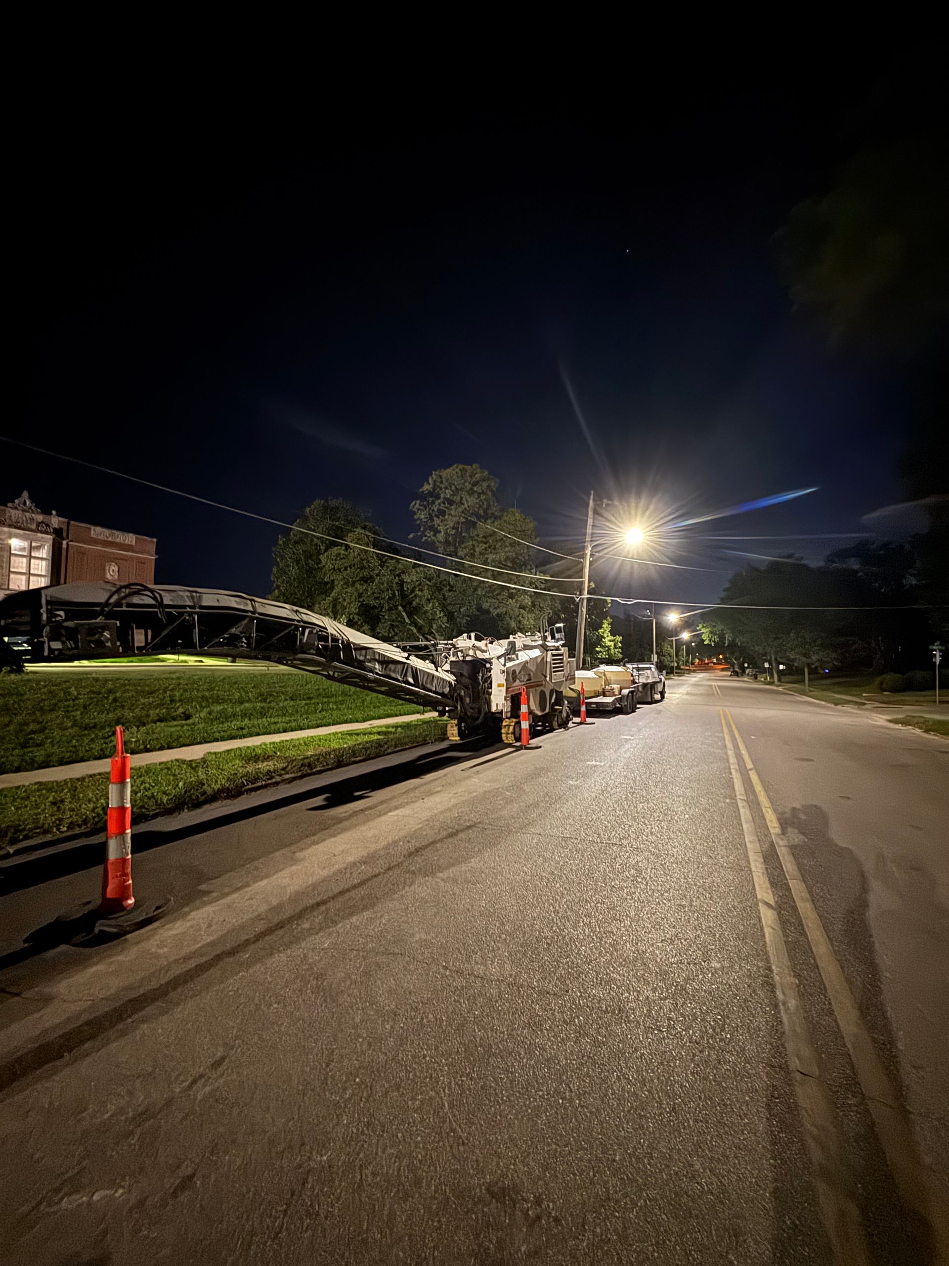 A nighttime view of a road milling machine parked on the side of a street next to a grassy curb with orange safety cones.