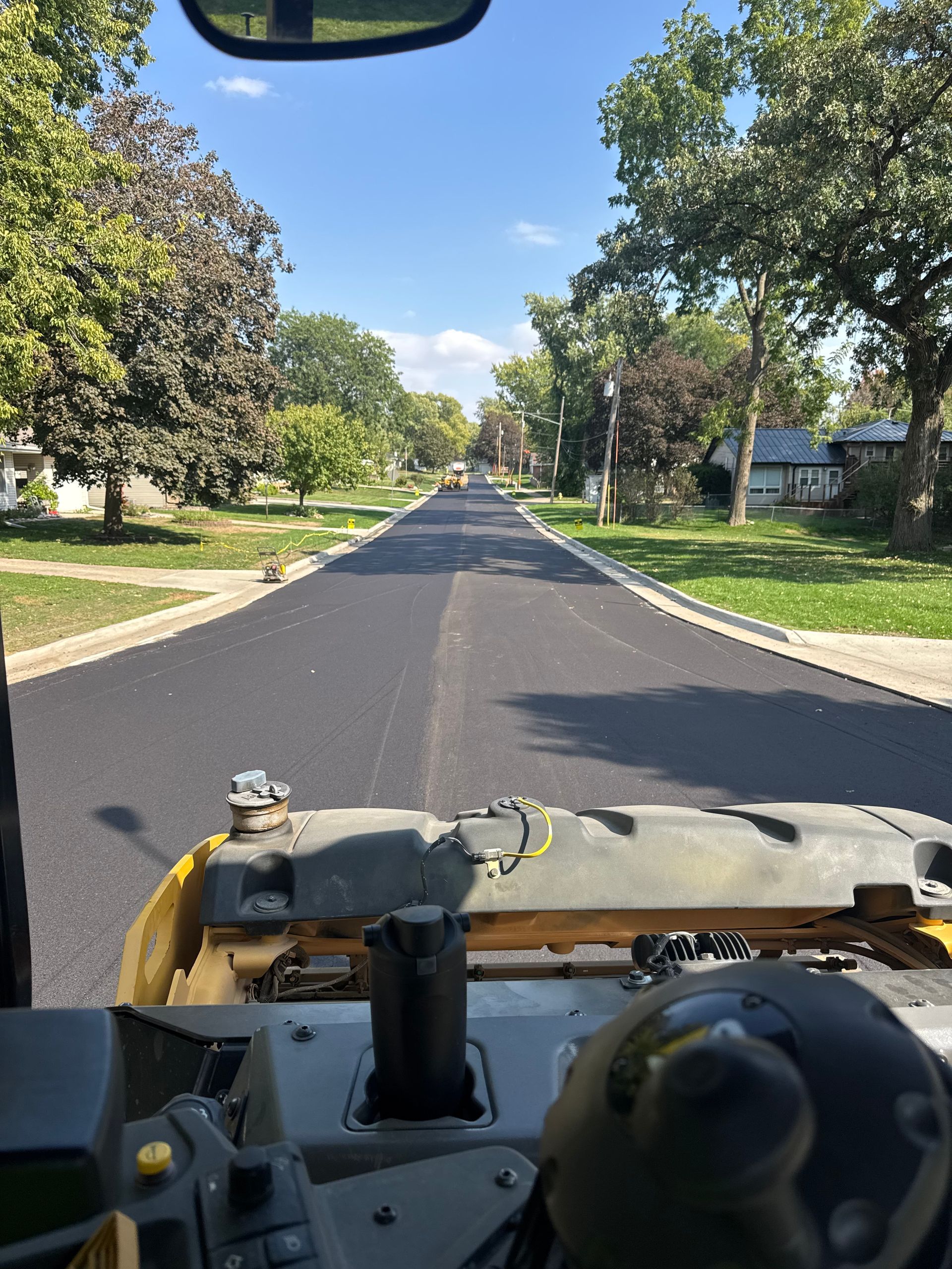 View from the driver's seat of construction equipment looking down a freshly paved, dark asphalt residential street.