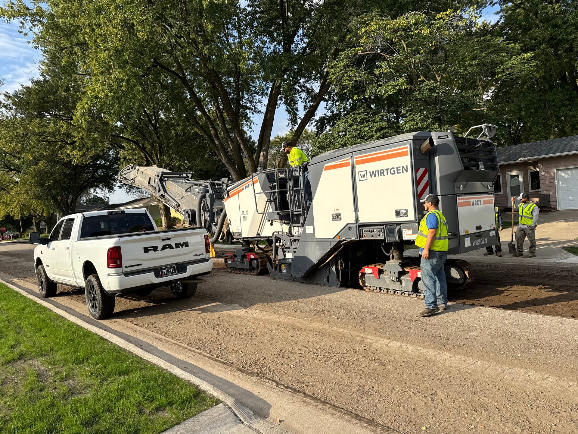 A white Ram pickup truck parked beside a large Wirtgen road milling machine on a gravel residential street.