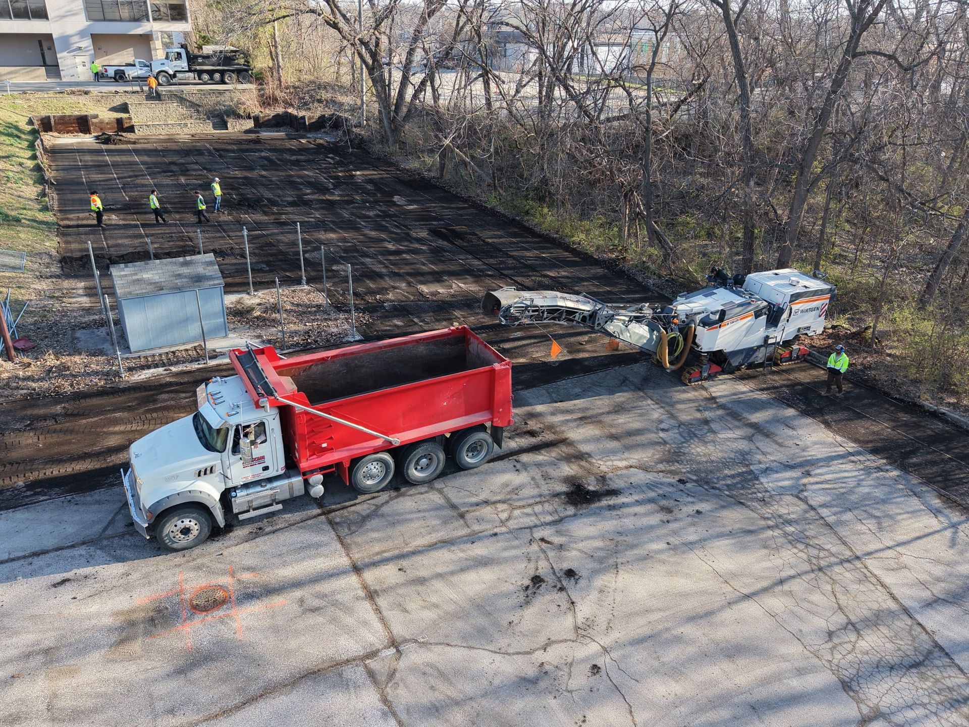 An aerial view shows a red dump truck beside a road milling machine on a construction site near a tree line.