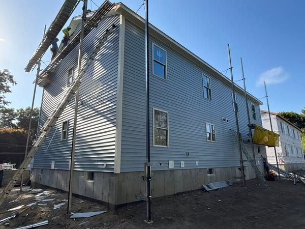 Construction of a two-story house with gray siding. Scaffolding, workers, and materials are present.