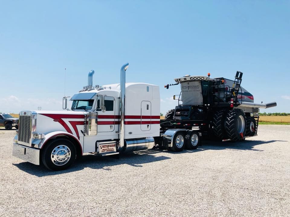 A semi truck is towing a tractor in a gravel lot.