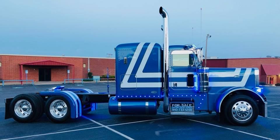 A blue semi truck is parked in a parking lot in front of a building.