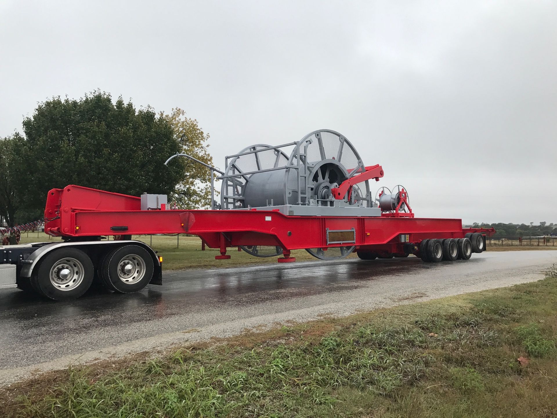 A red semi truck is carrying a large machine down a road.