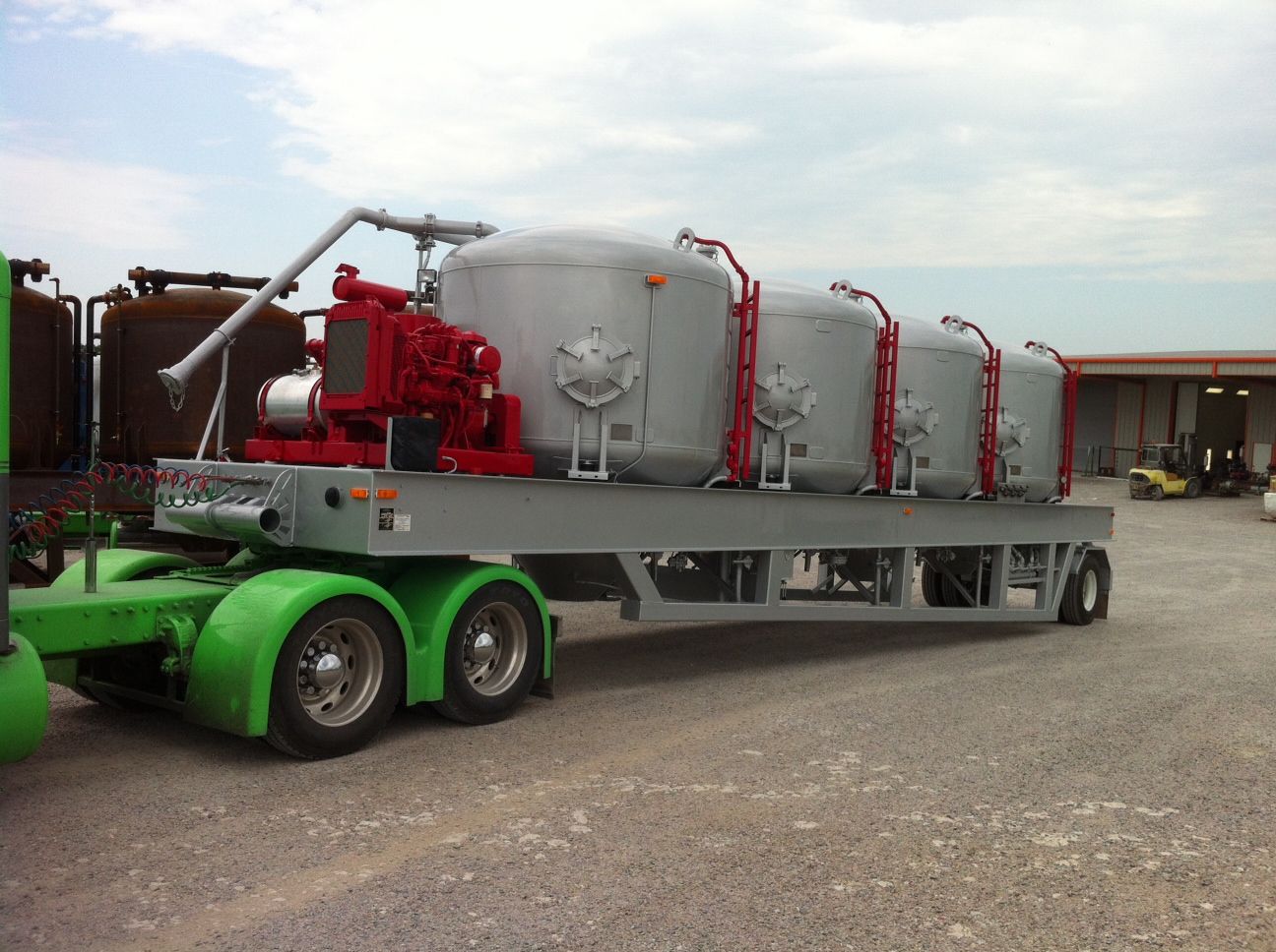 A green semi truck with tanks on the back is parked in a gravel lot
