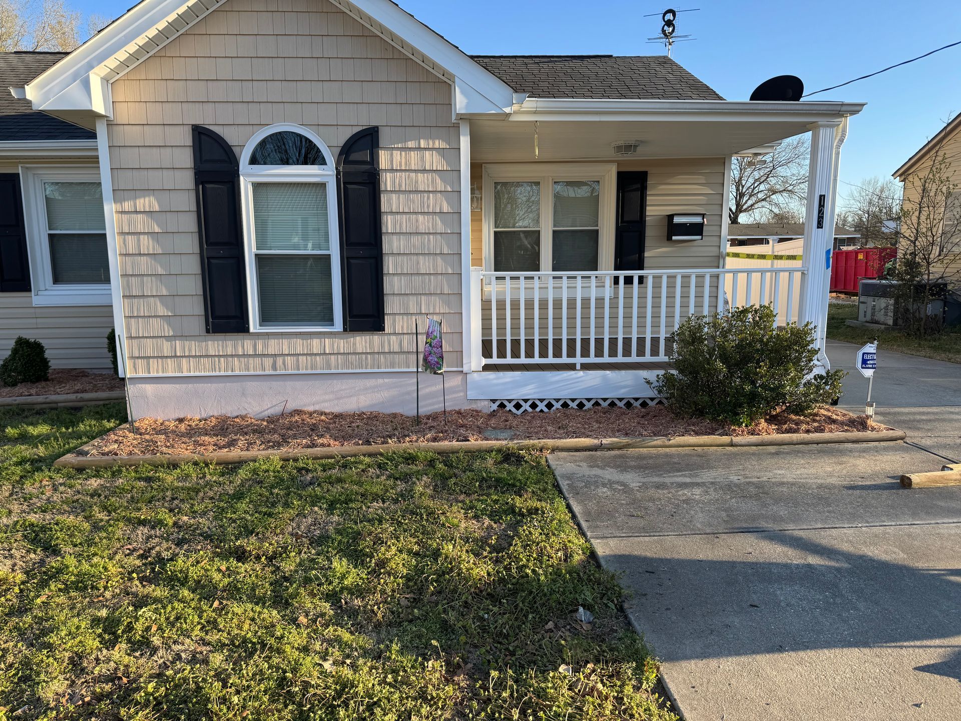 Small beige house with a porch and black shutters, set in a yard with a sidewalk