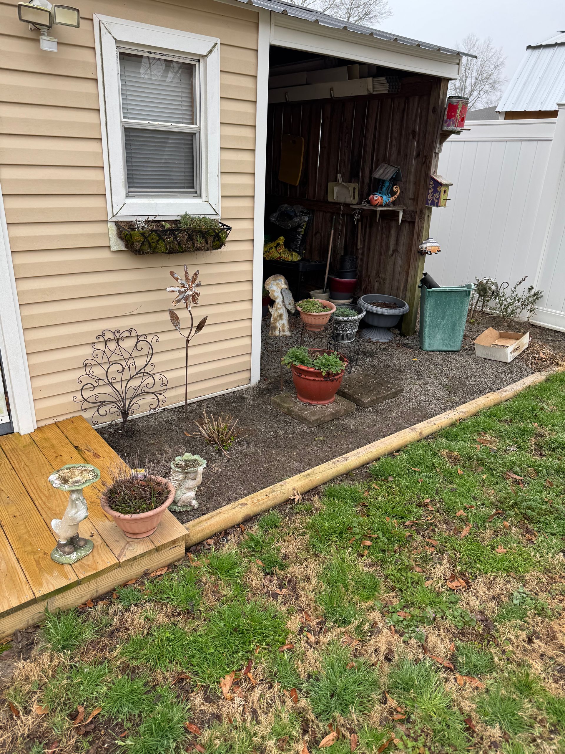 Tan shed with a small deck, window box, and a garden with various potted plants along the edge