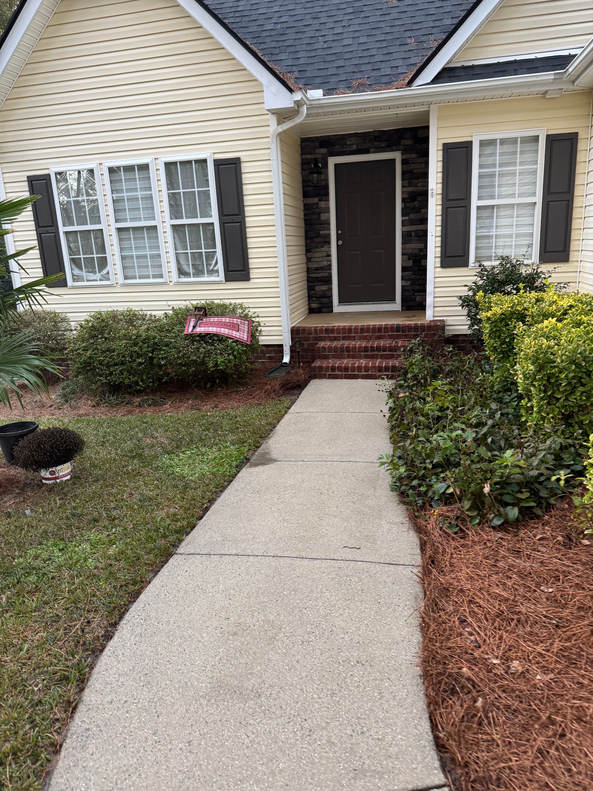 Sidewalk leading to a beige house with a dark brown door, dark shutters, and shrubs
