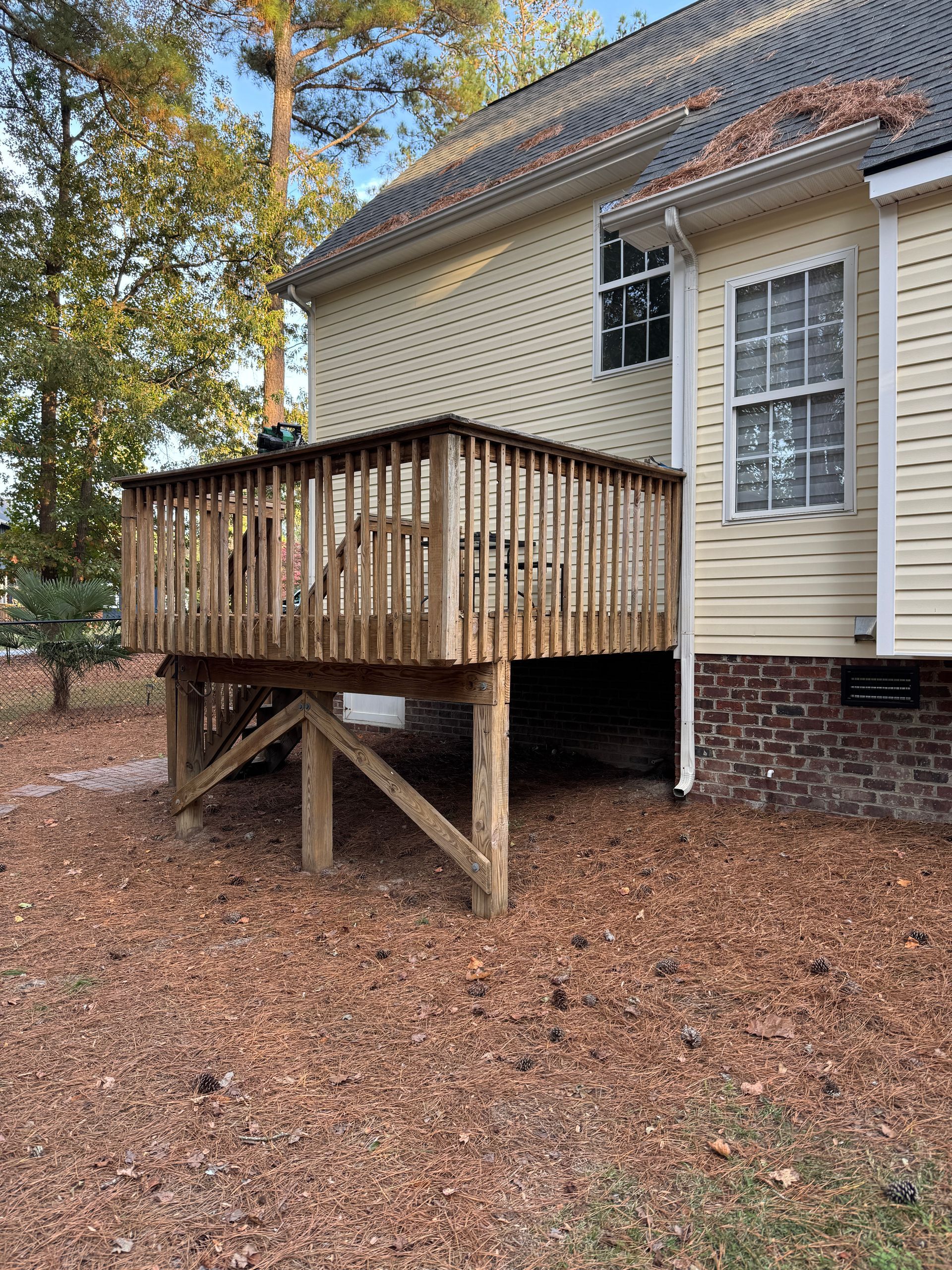 Wooden deck attached to a light yellow house with brick foundation, surrounded by fallen leaves