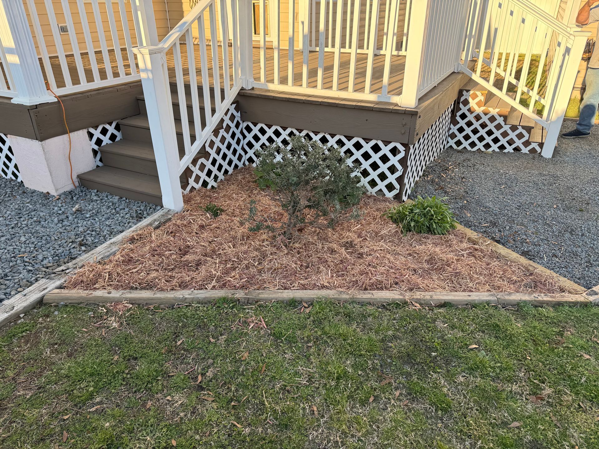 Triangular flower bed with wood mulch, a small bush, and a house porch with stairs