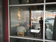 A person sits inside a windowed cafe, view through glass paneling. Outdoor seating is visible.
