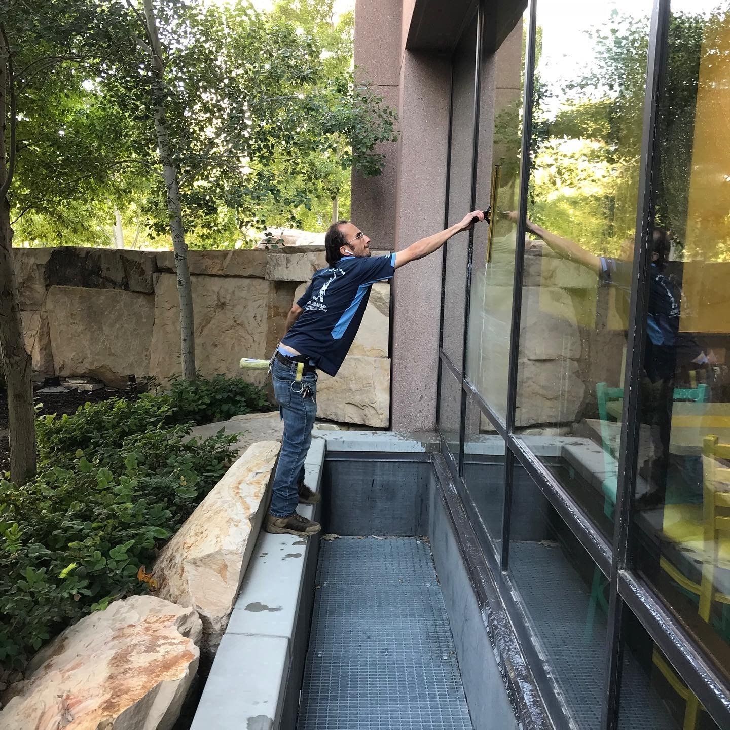A person cleaning a tall window outside. They're on a ledge with a metal grate below. Trees and stonework are visible.