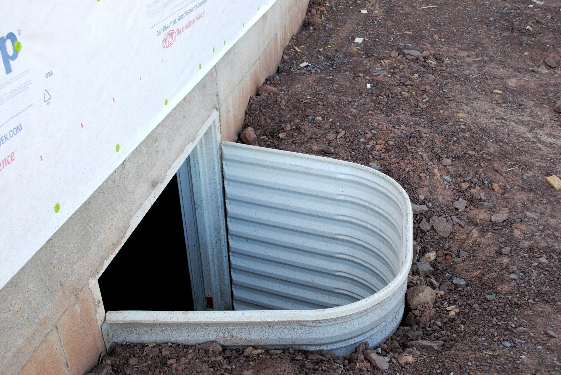 Window well with corrugated metal, in an unfinished basement setting. Soil surrounds the well.