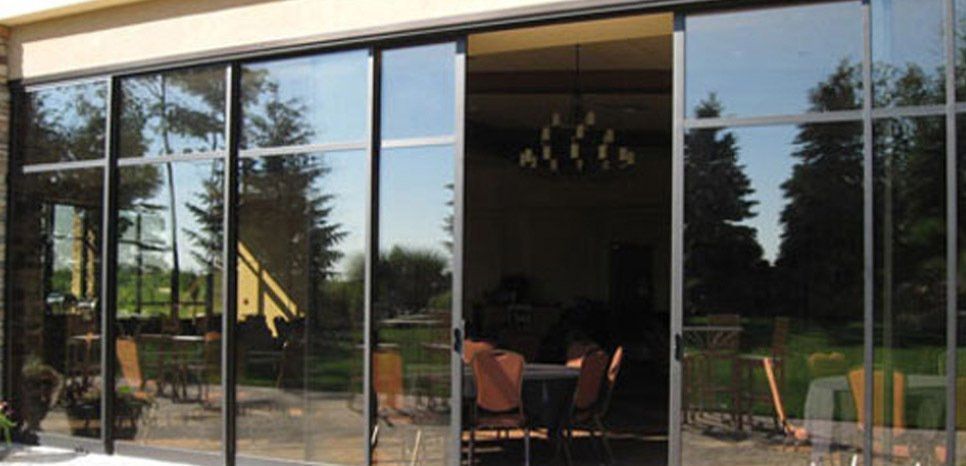 Sliding glass doors reflecting trees and sky, leading to an interior with a chandelier and table.