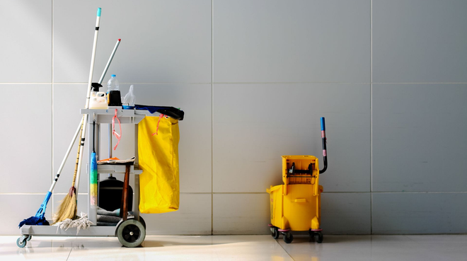 Cleaning supplies on carts: mops, brooms, buckets, and cleaning solutions against a tiled wall.