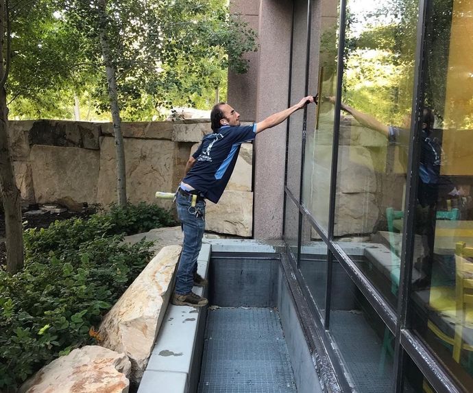 Man cleaning a large window with a squeegee outdoors. He is standing on a ledge next to a sunken window well.
