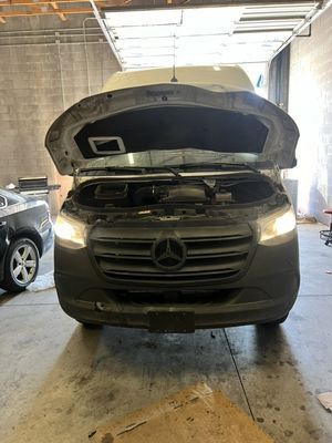 Mercedes-Benz Sprinter van with its hood open in a repair shop. Headlights are on.