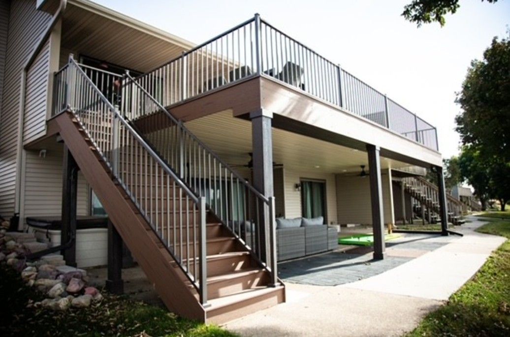 Exterior of a raised house with a wooden staircase, balcony, and covered patio in daylight