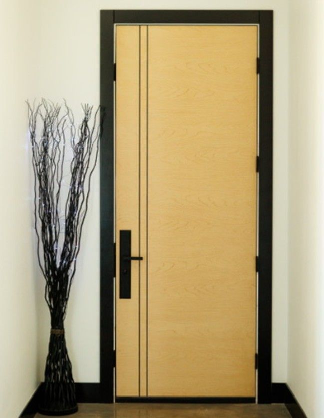 Closed light-wood door with black frame and handle beside tall black decorative branches