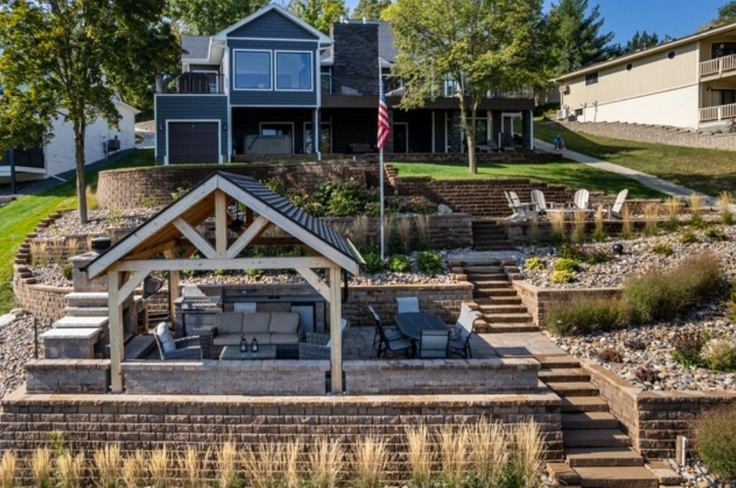 Tiered stone patio with a small pavilion and blue house above, overlooking a landscaped hillside and American flag