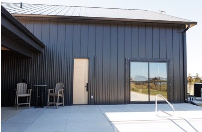 Modern dark-gray building exterior with patio chairs, a door, and a large window by a pool