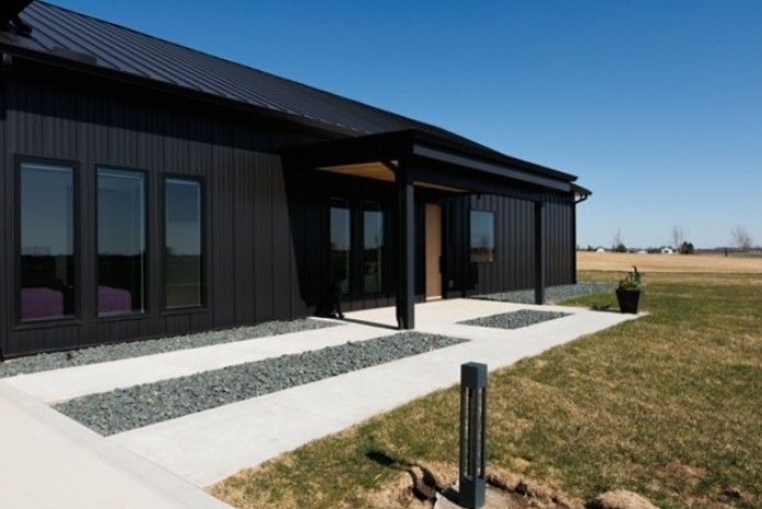 Modern black house with large windows and a concrete walkway in a grassy field under a blue sky