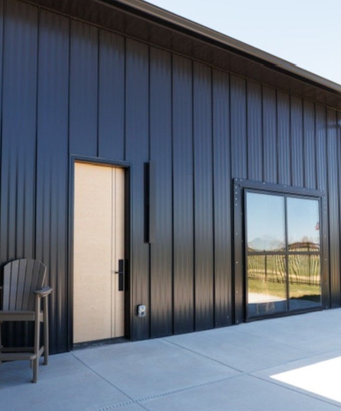 Black metal building exterior with a beige door, large window, and concrete patio in bright sunlight