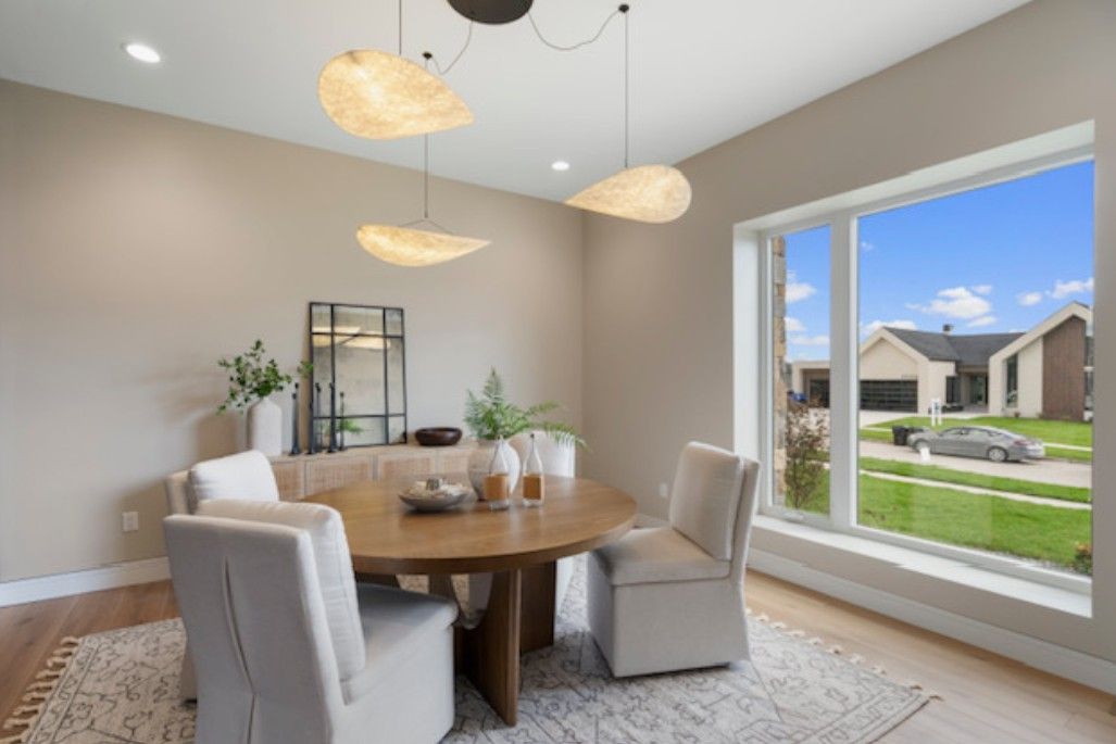 Bright dining room with round wood table, cream chairs, pendant lights, and a large window to a suburban street