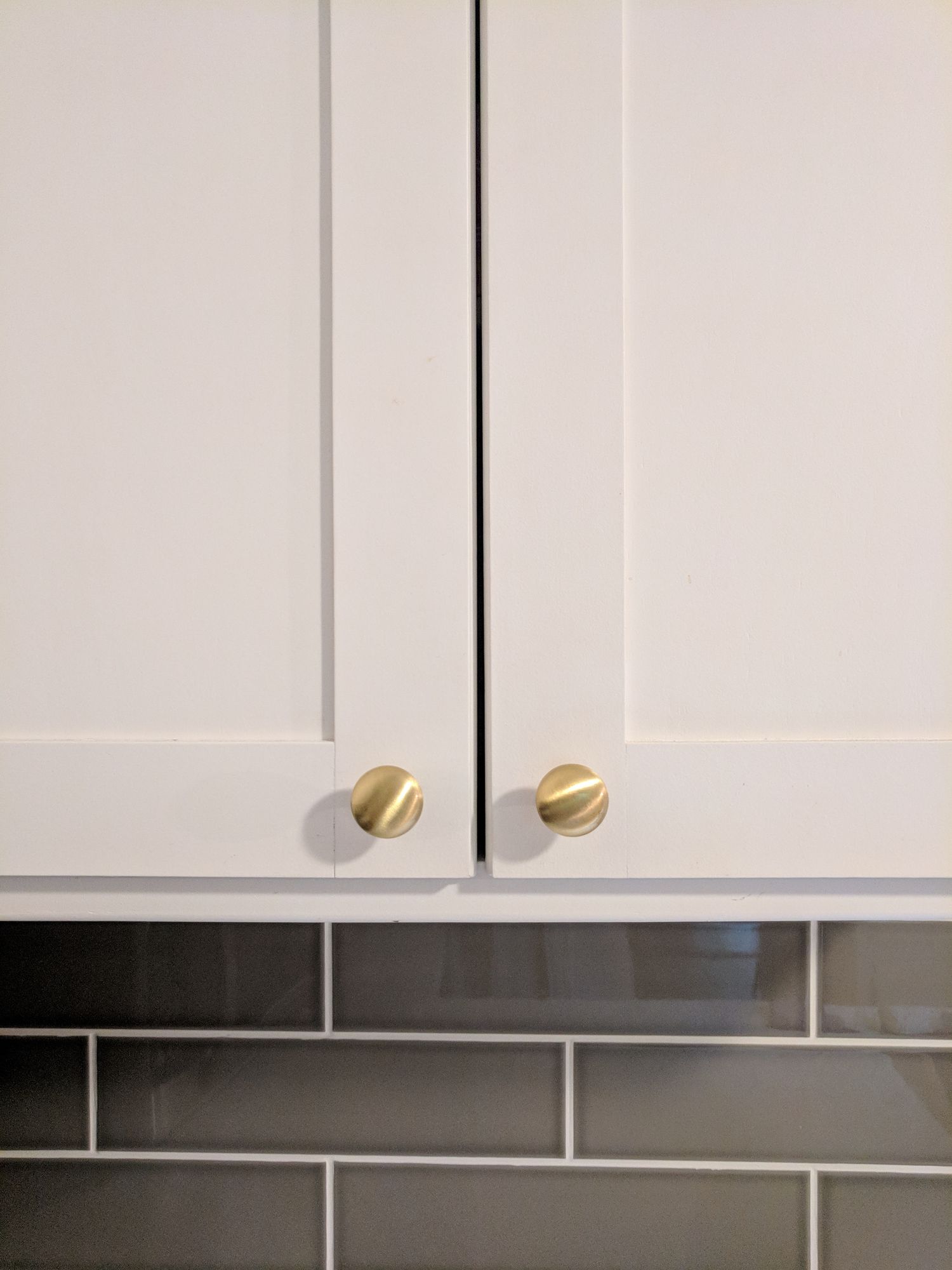 Two white shaker-style cabinet doors with round, brushed gold knobs, positioned above a grey subway tile backsplash.